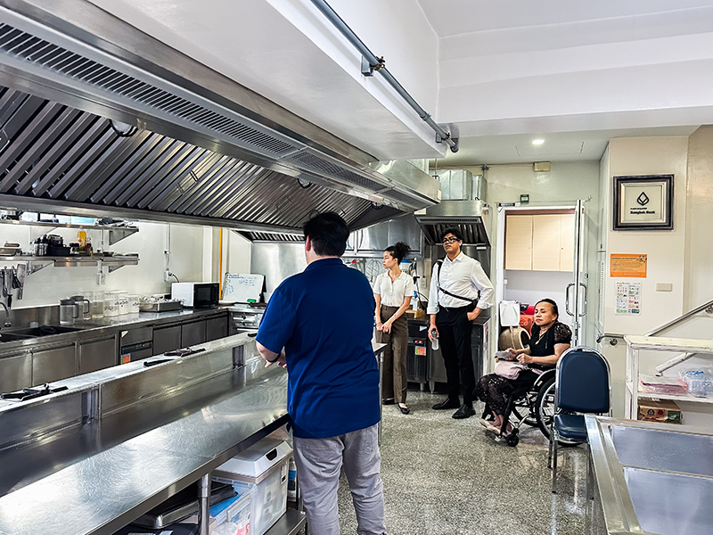 Picture 4: Mr. Pavarut Rungarunsunthon, Public Relations Officer (Center, facing away from the camera) provides information about kitchen for vocational training to Ms. Isabella Gacono; Mr. Krittapol Chamrasromran; and Ms. Nongnuch Maytarjittipun (seated in wheelchair) in the background.