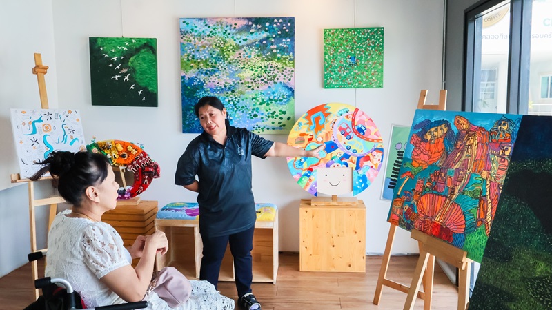 Picture 9: Ms. Nongnuch Maytarjittipun (left, seated in a wheelchair) listening to staff providing information art works made by persons with autism