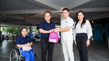 Picture 1: Participants pose for a group photo. Mr. Piroon Laismit and Mr. Asdang Sukwises hold souvenirs, while Ms. Nongnuch Maytarjittipun is seated in a wheelchair on the left side of the group